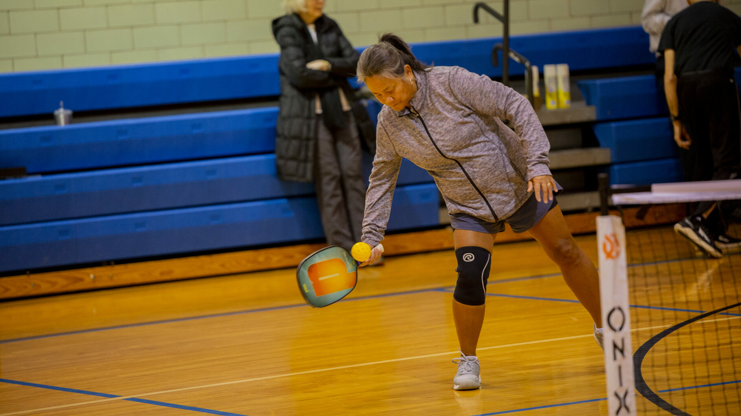 Woman in grey sweatshirt and shorts leaning down to hit the ball in a picklleball game.