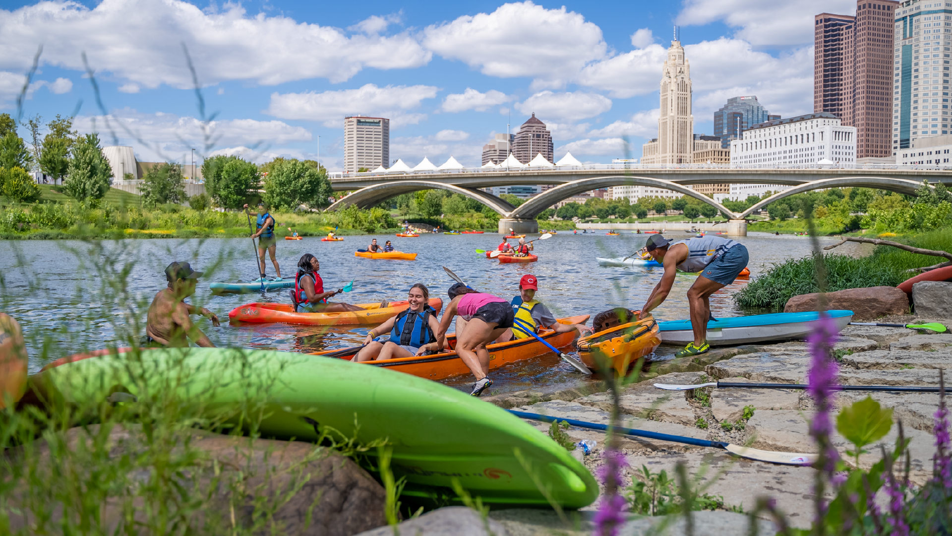 A group of people enjoying the Scioto River in kayaks with the downtown Columbus skyline in the background.